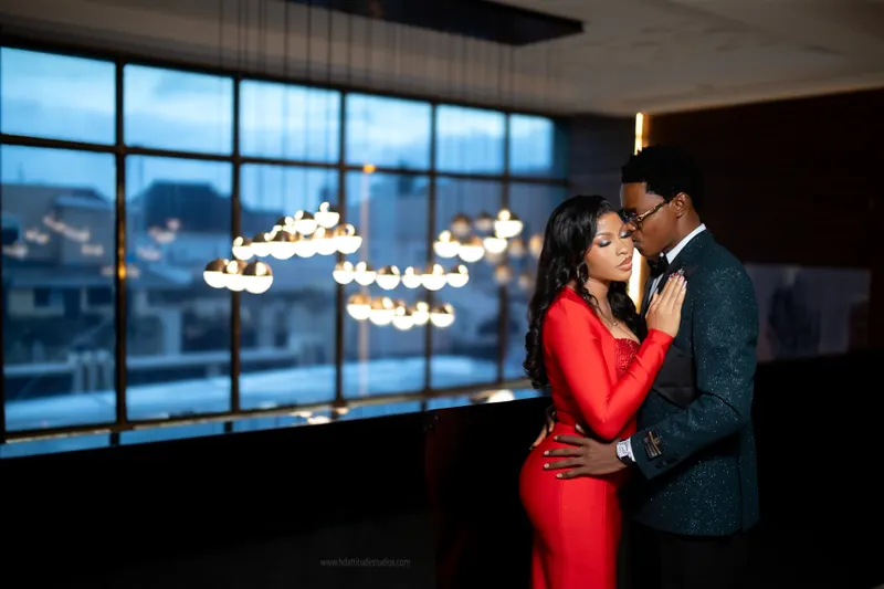 Couple embracing beside floor-to-ceiling window at dusk, cluster of round pendant lights, city skyline visible