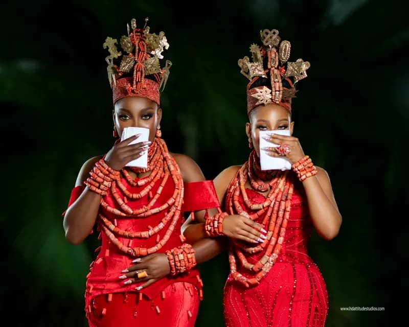 Two women in matching coral-bead crowns and red gowns, each holding white cloth to lips, dark green foliage backdrop